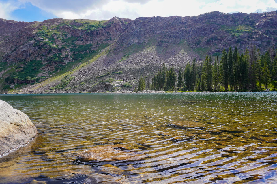 The image depicts a serene mountain lake surrounded by lush green trees and shrubs, with a rocky cliff face on one side. The calm water surface reflects the clear blue sky above, creating a tranquil atmosphere. The perspective is from the shoreline, looking out towards the mountains in the background.
