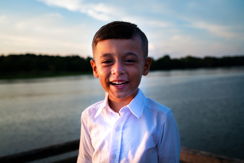 Niño sonriente en camisa de vestir blanca de pie cerca del cuerpo de agua durante el día