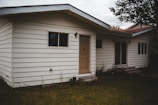 A one-story house with white horizontal siding, and a beige front door. There are two windows on either side of the door, and two wall-mounted light fixtures next to the entrance. The house is surrounded by a grassy area with some overgrown vegetation near the walls and a tree branch visible at the top right corner.