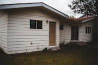A one-story house with white horizontal siding, and a beige front door. There are two windows on either side of the door, and two wall-mounted light fixtures next to the entrance. The house is surrounded by a grassy area with some overgrown vegetation near the walls and a tree branch visible at the top right corner.