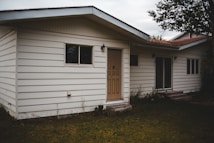 A one-story house with white horizontal siding, and a beige front door. There are two windows on either side of the door, and two wall-mounted light fixtures next to the entrance. The house is surrounded by a grassy area with some overgrown vegetation near the walls and a tree branch visible at the top right corner.