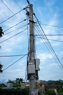A utility pole with numerous electrical wires and cables attached, set against a partly cloudy sky. The pole is situated near some buildings and trees, contributing to a suburban or rural setting.