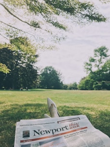 A person is lying on grass with crossed legs holding a newspaper. The scene is set in a park with lush greenery and a tree canopy overhead, giving a peaceful, leisurely atmosphere.