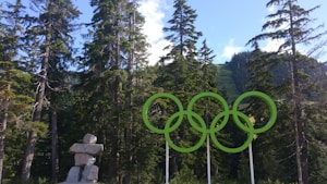 Green Olympic rings are prominently displayed against a backdrop of tall evergreen trees and a clear blue sky. A stone inukshuk is present on the left side of the image.