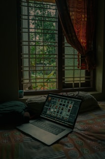 A room with a soft, cozy atmosphere features a laptop open on a bed. The room is dimly lit with natural light filtering through a window with bars. Outside, green trees can be seen, adding a refreshing touch to the scene. A patterned curtain partially covers the window.