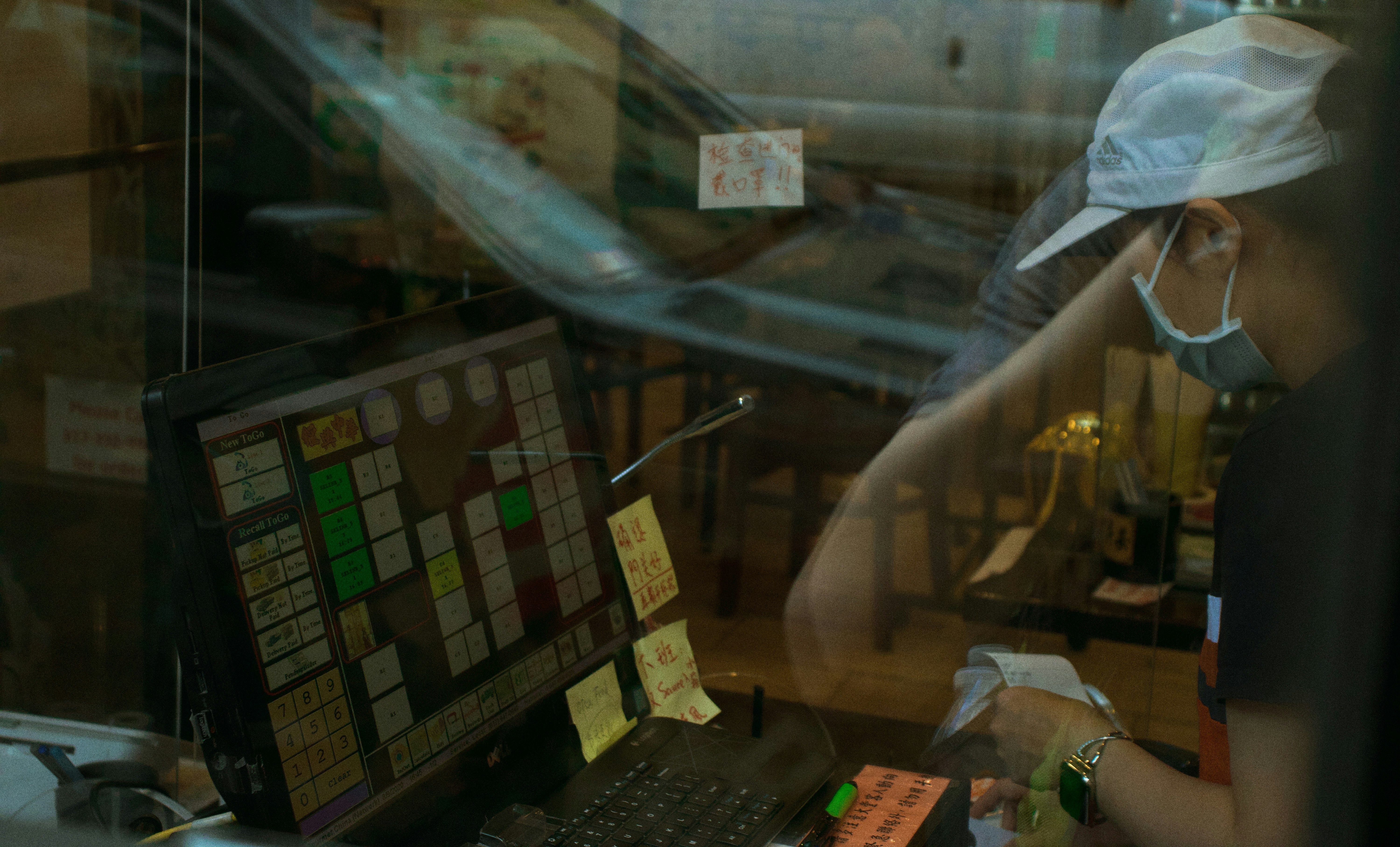 A person wearing a cap, face mask, and dark clothes interacts with a point-of-sale system at a counter. The image includes reflections, giving a sense of separation by glass. Sticky notes and written messages are visible near the screen and on the counter. The background seems to show a somewhat cluttered interior with chairs and tables.