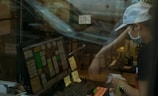 A person wearing a cap, face mask, and dark clothes interacts with a point-of-sale system at a counter. The image includes reflections, giving a sense of separation by glass. Sticky notes and written messages are visible near the screen and on the counter. The background seems to show a somewhat cluttered interior with chairs and tables.