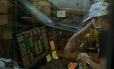 A person wearing a cap, face mask, and dark clothes interacts with a point-of-sale system at a counter. The image includes reflections, giving a sense of separation by glass. Sticky notes and written messages are visible near the screen and on the counter. The background seems to show a somewhat cluttered interior with chairs and tables.