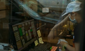 A person wearing a cap, face mask, and dark clothes interacts with a point-of-sale system at a counter. The image includes reflections, giving a sense of separation by glass. Sticky notes and written messages are visible near the screen and on the counter. The background seems to show a somewhat cluttered interior with chairs and tables.