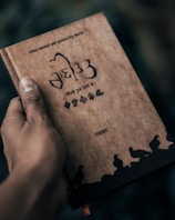 A hand is holding a book with a brown cover. The book has text and decorative elements in a script that appears to be Punjabi or Hindi. The cover features illustrations of birds along the bottom and various floral patterns. The background is blurred and dark.