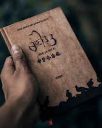 A hand is holding a book with a brown cover. The book has text and decorative elements in a script that appears to be Punjabi or Hindi. The cover features illustrations of birds along the bottom and various floral patterns. The background is blurred and dark.