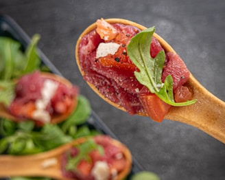 A wooden spoon holds a serving of sliced raw tuna, garnished with arugula and diced tomatoes, sprinkled with coarse salt and black pepper. The focus is on the fresh ingredients, with additional spoons blurred in the background.