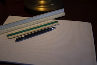 A close-up view of graph paper placed on a dark surface, with a black mechanical pencil and two rulers lying on top. The rulers have measurement markings and are positioned perpendicular to each other. A table lamp base is visible in the background.