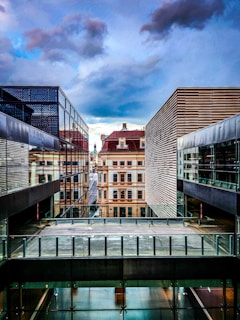 A modern architectural scene with two contemporary buildings featuring glass and metal facades. In the background, an older, classical building adds contrast with its ornate design. The sky is overcast with dramatic clouds, creating a sense of depth and drama.