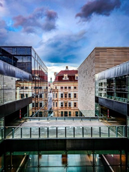 A modern architectural scene with two contemporary buildings featuring glass and metal facades. In the background, an older, classical building adds contrast with its ornate design. The sky is overcast with dramatic clouds, creating a sense of depth and drama.