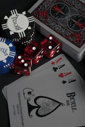 A collection of poker chips, red dice, and a deck of playing cards is arranged on a dark surface. The cards include an ace of spades with intricate black artwork and a standard Bicycle design. The set includes black, white, and blue poker chips featuring the Bicycle brand name.