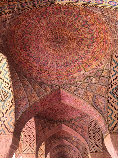 A ceiling adorned with intricate and colorful Persian patterns, featuring floral and geometric motifs. The arches are decorated with similar detailed designs, reflecting traditional Persian architecture.