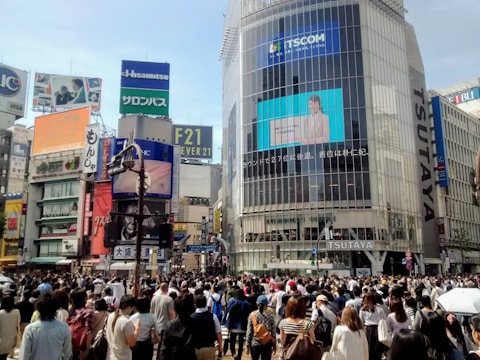 A bustling urban scene featuring a large crowd of people crossing a busy intersection surrounded by tall buildings covered with advertisements and signage, including a prominent screen displaying a video on the side of a building.