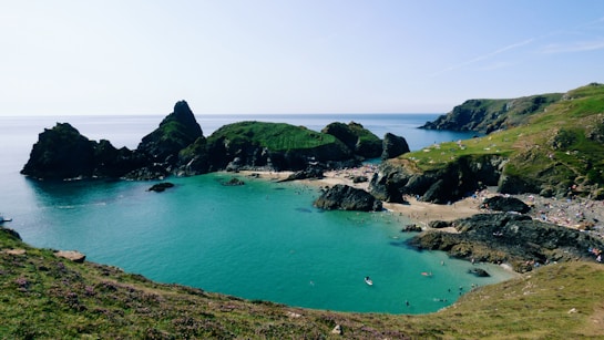 A scenic coastal landscape featuring a turquoise bay surrounded by rugged cliffs and rocky formations. The beach area is populated with people enjoying the sun and sea. The lush greenery on the cliffs contrasts with the deep blue of the sea under the clear sky.