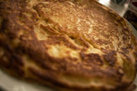 A close-up view of a golden-brown pancake with a crispy texture. The pancake appears to be freshly cooked, showing a mix of darker and lighter brown patches across its surface. The soft, fluffy interior is slightly visible, and the pancake is placed on a light-colored plate.
