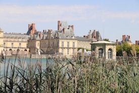 A historic chateau with ornate architecture stands by a calm lake, surrounded by lush greenery and reeds in the foreground. The building features numerous chimneys and arched windows, highlighted by clear blue skies in the background.