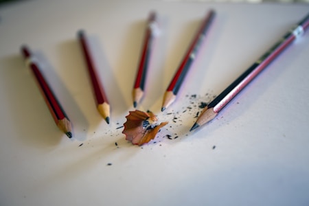 Five sharpened pencils are arranged on a white surface, with shavings and pencil lead scattered around them. The pencils have a red and black exterior with a wooden tip exposed. One pencil appears to be freshly sharpened, with shavings prominently displayed.