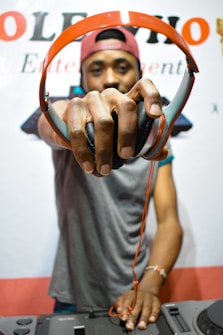 A person wearing a red cap holds out a pair of red and black headphones towards the camera, with a DJ mixer in the foreground.
