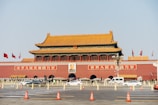 A large, traditional Chinese gate with a golden roof and red walls, featuring multiple flags. A prominent portrait is displayed above the central entrance, with vehicles and pedestrians passing in front.