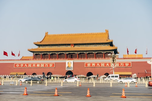 A large, traditional Chinese gate with a golden roof and red walls, featuring multiple flags. A prominent portrait is displayed above the central entrance, with vehicles and pedestrians passing in front.