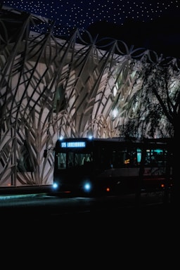 A bus with the destination 'Luxembourg' is illuminated by its headlights at night. It is positioned in front of a modern, intricately designed building with a unique architectural pattern. The sky is dark, dotted with many small, bright stars, enhancing the nighttime atmosphere.