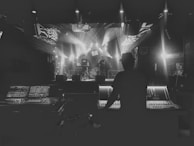 A person is operating a large sound mixing console, facing a stage where musicians are performing. The stage is illuminated by bright spotlights, creating a dramatic and moody atmosphere. The venue appears to be a dimly lit music hall or club.