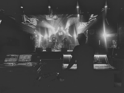 A person is operating a large sound mixing console, facing a stage where musicians are performing. The stage is illuminated by bright spotlights, creating a dramatic and moody atmosphere. The venue appears to be a dimly lit music hall or club.