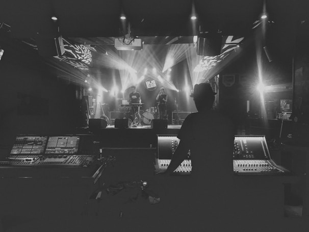 A person is operating a large sound mixing console, facing a stage where musicians are performing. The stage is illuminated by bright spotlights, creating a dramatic and moody atmosphere. The venue appears to be a dimly lit music hall or club.