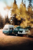 Two vintage camper vans are parked on a grassy area surrounded by tall pine trees. The vans have unique designs, with one colored blue and white and the other green with geometric mountain graphics. Sunlight filters through the trees, creating a warm and inviting atmosphere. The scene captures a sense of adventure and freedom in a natural setting.