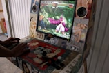 An arcade machine with a retro design features a fighting game on the screen. A person is using the joystick and buttons to play. The screen displays two characters in a battle scene set in a forest environment. Speakers are mounted on either side of the screen, and the arcade cabinet is decorated with comic-style art and pin-up illustrations.