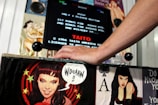 A hand is resting on the control panel of an arcade machine, which is adorned with pin-up style illustrations of women and comic-style speech. The screen displays text from an arcade game and includes the name TAITO and the year 1986.