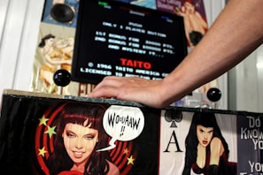 A hand is resting on the control panel of an arcade machine, which is adorned with pin-up style illustrations of women and comic-style speech. The screen displays text from an arcade game and includes the name TAITO and the year 1986.