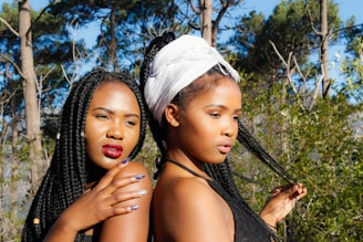 Two women are standing closely in a natural outdoor setting with trees and blue sky in the background. Both have long braided hair, and one of them is wearing a white headscarf. They appear to be posing confidently with one looking directly ahead and the other looking off to the side, showcasing bold makeup and stylish attire.