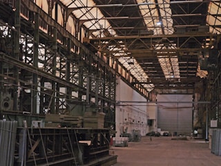 A large, empty industrial warehouse with high ceilings and metal support structures. The floor is bare, with scattered equipment and materials. The walls are lined with metal beams and there are a few vehicles parked in the background.
