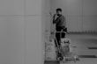A person wearing a mask is standing in an indoor setting holding a mop. The individual appears to be engaged in cleaning activities, accompanied by a cleaning cart filled with supplies. Caution signs are visible, indicating a wet floor. The environment is spacious and tiled, with a partially visible wall in the background.