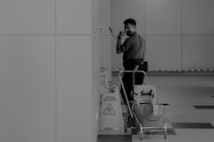 A person wearing a mask is standing in an indoor setting holding a mop. The individual appears to be engaged in cleaning activities, accompanied by a cleaning cart filled with supplies. Caution signs are visible, indicating a wet floor. The environment is spacious and tiled, with a partially visible wall in the background.
