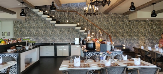 A well-lit and stylish dining area features an elegant, rustic design with a stone accent wall, exposed wooden beams, and black chandeliers hanging from the ceiling. The room is furnished with neatly arranged tables set with white napkins, plates, and glasses, while the back section includes a counter with kitchenwares and a variety of food items displayed, likely for a buffet setting. Stairs ascend along the stone wall, adding architectural interest.