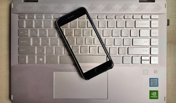 A smartphone is placed on top of a laptop keyboard. The keyboard is part of a metallic laptop, and the smartphone is positioned diagonally across the keys. The laptop features visible logos near the bottom right corner, including Intel and NVIDIA stickers, indicating the hardware specifications.