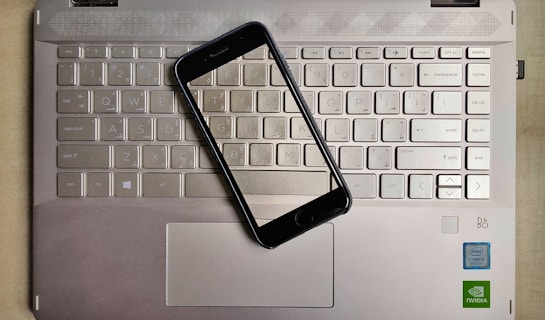 A smartphone is placed on top of a laptop keyboard. The keyboard is part of a metallic laptop, and the smartphone is positioned diagonally across the keys. The laptop features visible logos near the bottom right corner, including Intel and NVIDIA stickers, indicating the hardware specifications.