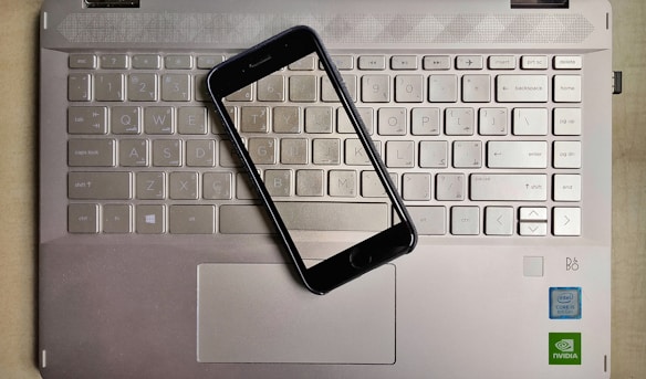 A smartphone is placed on top of a laptop keyboard. The keyboard is part of a metallic laptop, and the smartphone is positioned diagonally across the keys. The laptop features visible logos near the bottom right corner, including Intel and NVIDIA stickers, indicating the hardware specifications.