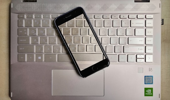 A smartphone is placed on top of a laptop keyboard. The keyboard is part of a metallic laptop, and the smartphone is positioned diagonally across the keys. The laptop features visible logos near the bottom right corner, including Intel and NVIDIA stickers, indicating the hardware specifications.