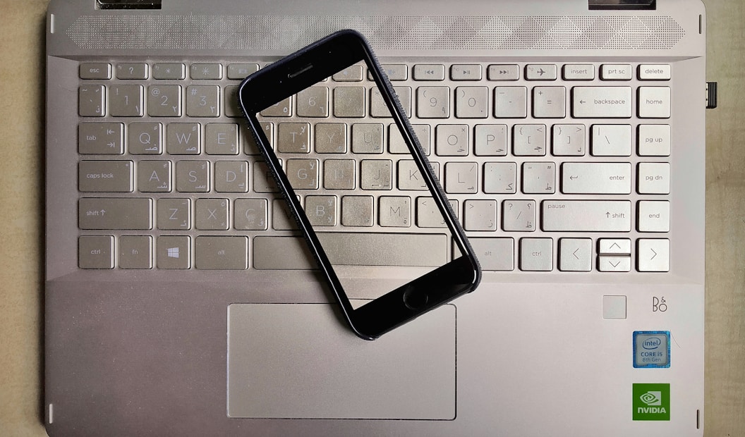 A smartphone is placed on top of a laptop keyboard. The keyboard is part of a metallic laptop, and the smartphone is positioned diagonally across the keys. The laptop features visible logos near the bottom right corner, including Intel and NVIDIA stickers, indicating the hardware specifications.