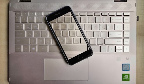 A smartphone is placed on top of a laptop keyboard. The keyboard is part of a metallic laptop, and the smartphone is positioned diagonally across the keys. The laptop features visible logos near the bottom right corner, including Intel and NVIDIA stickers, indicating the hardware specifications.