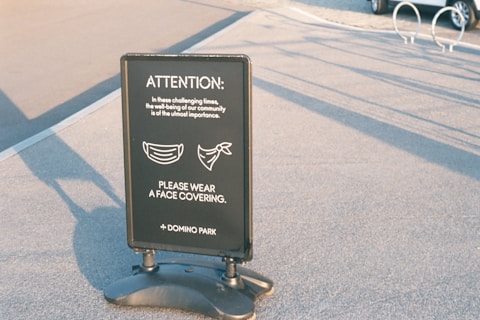A standing sign on a paved area displays a message regarding the importance of wearing face coverings for community well-being. The sign features images of two masks and is attributed to Domino Park.