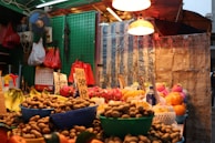 A vibrant market stall displays various fruits in baskets, including bananas, oranges, and dragon fruit. Handwritten price signs and protective nets for some fruits are visible. Red and orange hanging bags, blue waterproof tarp, and overhead lights illuminate the scene, creating a warm, bustling atmosphere.
